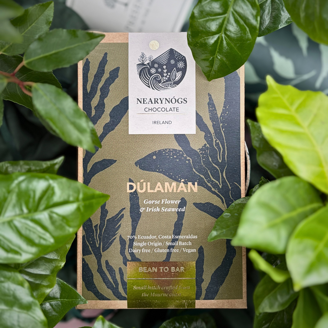 A NearyNógs Gorse Flower & Irish Seaweed chocolate bar with seaweed design and branding, nestled in amongst green leaves with The Present Tree tube in the background.